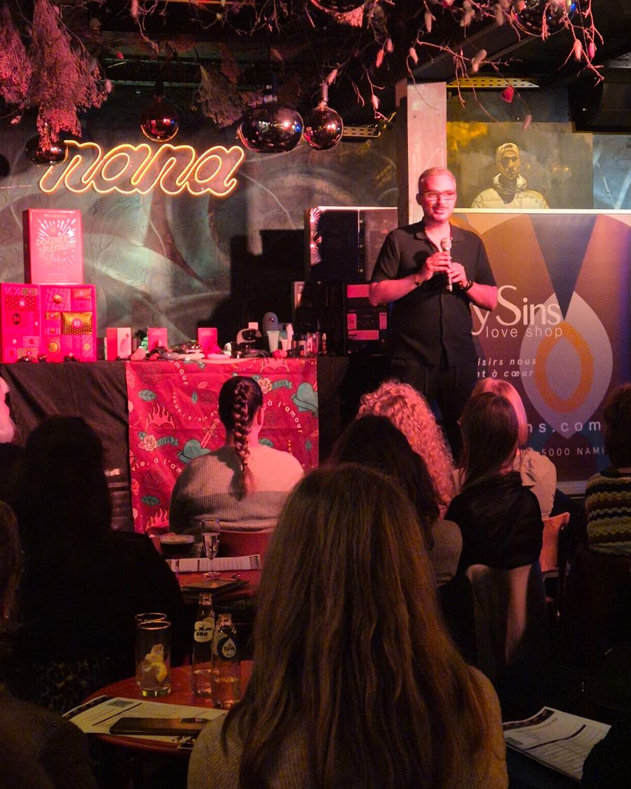 Une personne de l'équipe Lovely Sins au micro devant des calendriers de l'Avent et des produits exposés, avec le public au premier plan, lors des Lovely Talks à Namur.